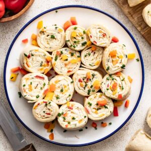 A plate of vegetable tortilla pinwheels with cream cheese and a bowl of peppers next to them.