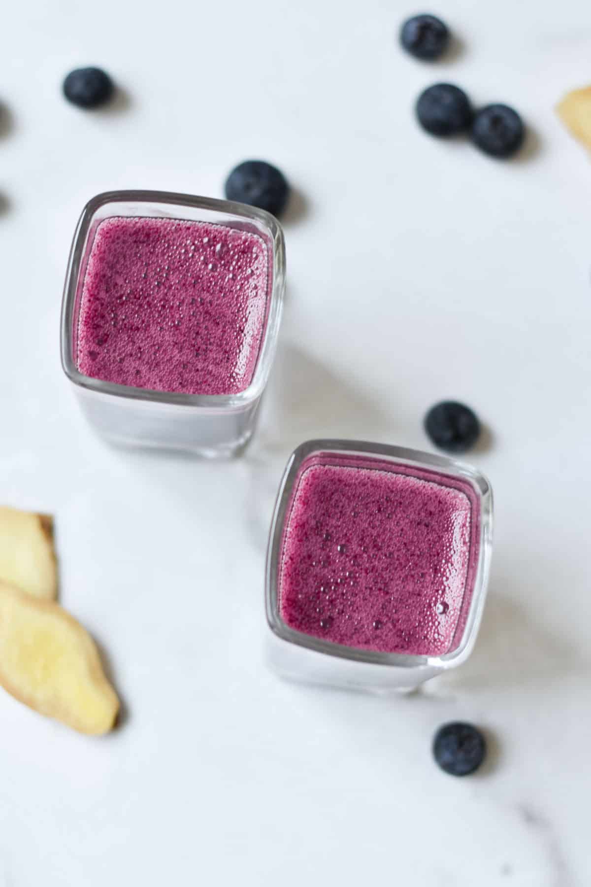 Two ginger blueberry shots on a white background with fresh blueberries and sliced ginger root.