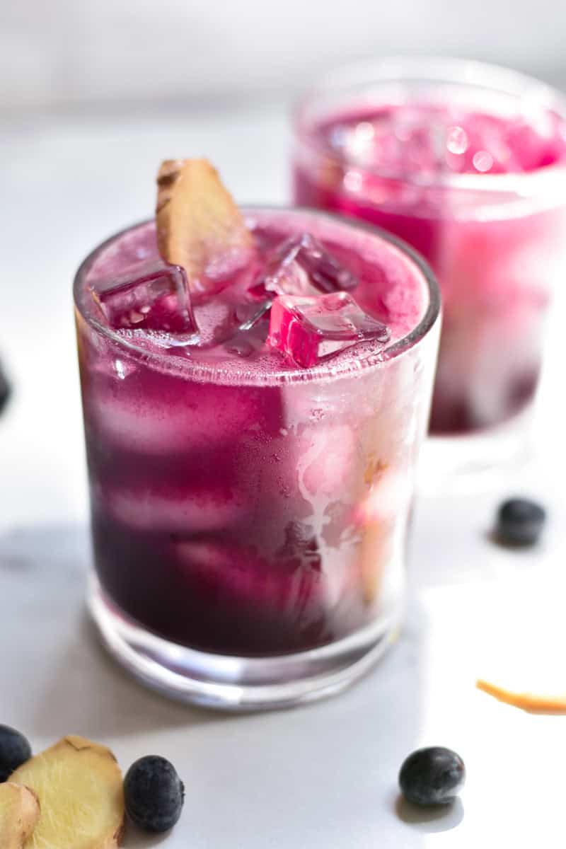 Two blueberry mocktails with ginger on a white table with fresh blueberries and ginger next to the glass.