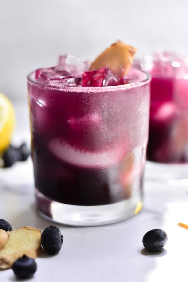 A bright blueberry drink with a slice of ginger in a short glass.