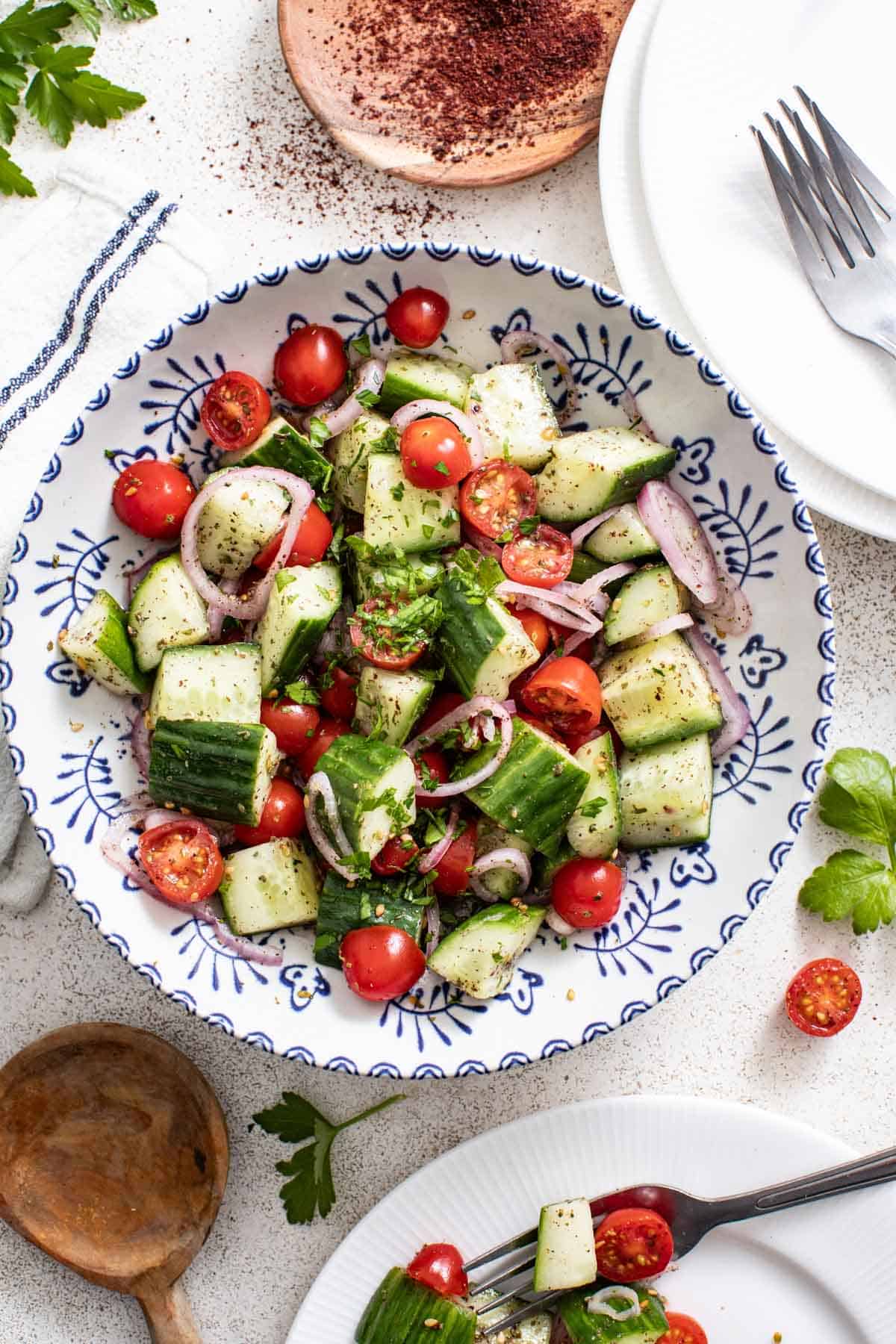 Cucumber and tomato salad in a white and blue bowl next to plates, a wood spoon, and a bowl of sumac.