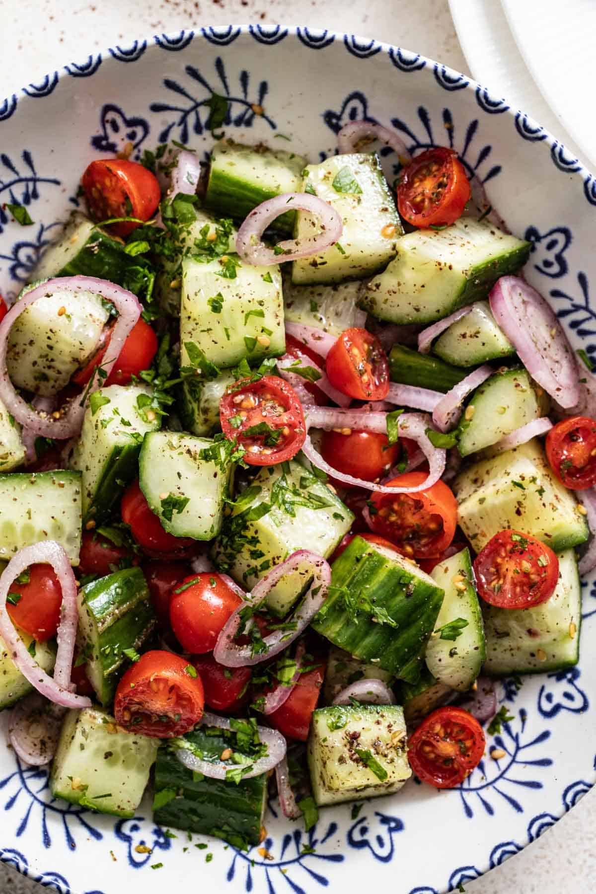 A blue and white patterned bowl filled with cucumber and tomato salad with mediterranean spices on top.