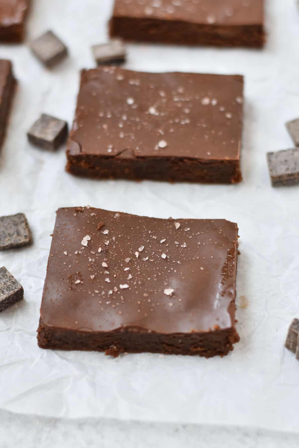 Gooey brownies lined up in a row on wax paper with chocolate pieces sprinkled around them.