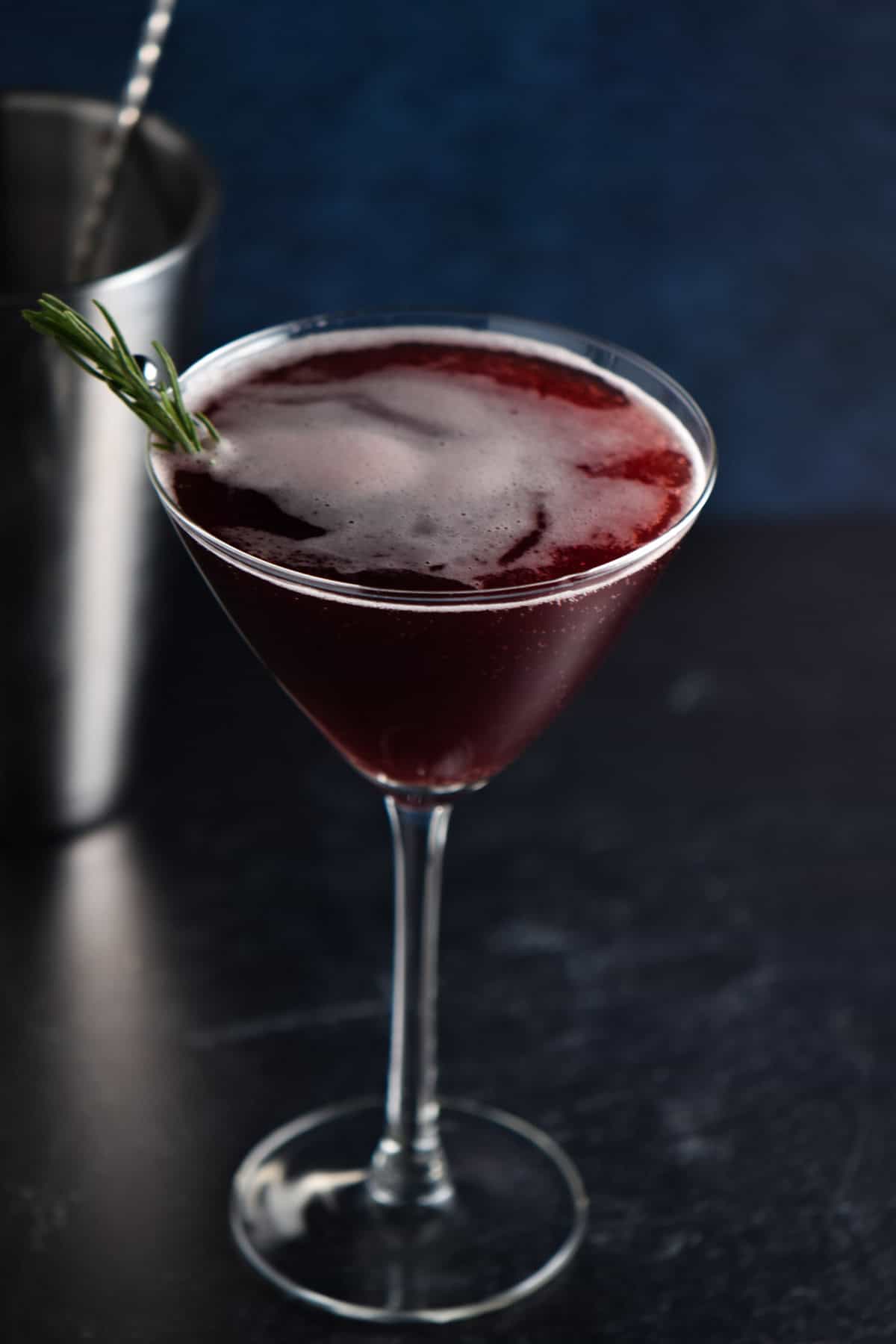 A mocktail with a sprig of rosemary on top in front of a silver shaker.