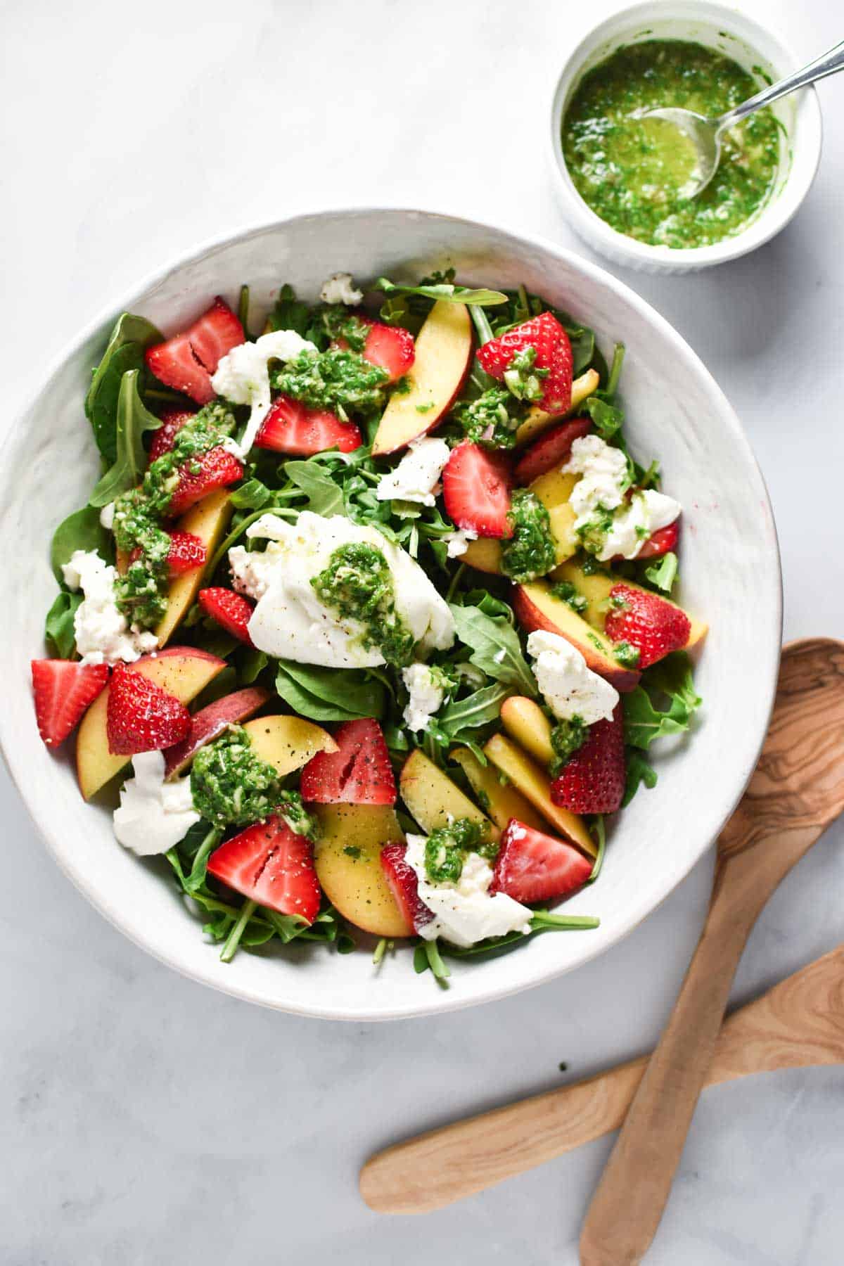 A bowl of burrata and peach salad next to a cup of vinaigrette and two wooden serving spoons.