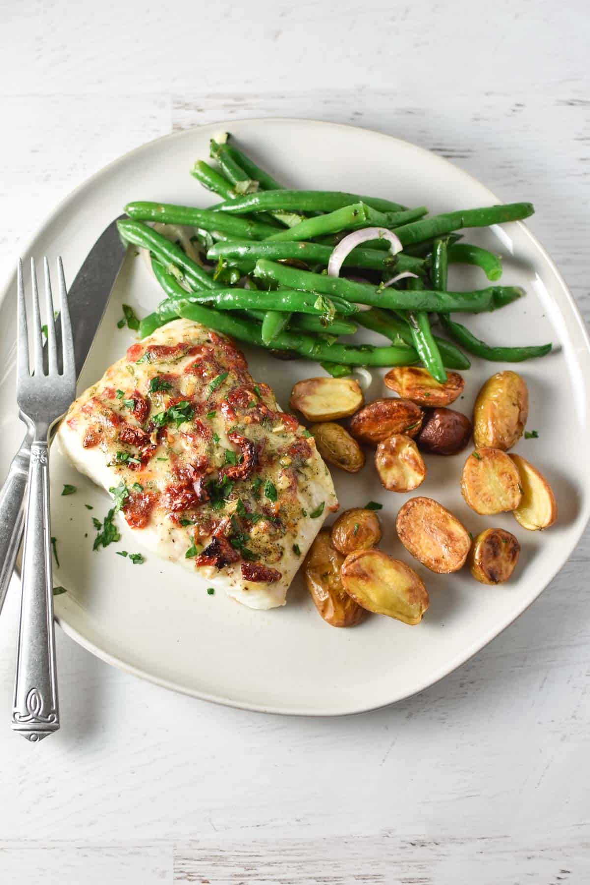 Mediterranean baked halibut on a plate with potatoes and green beans, next to a knife and fork.