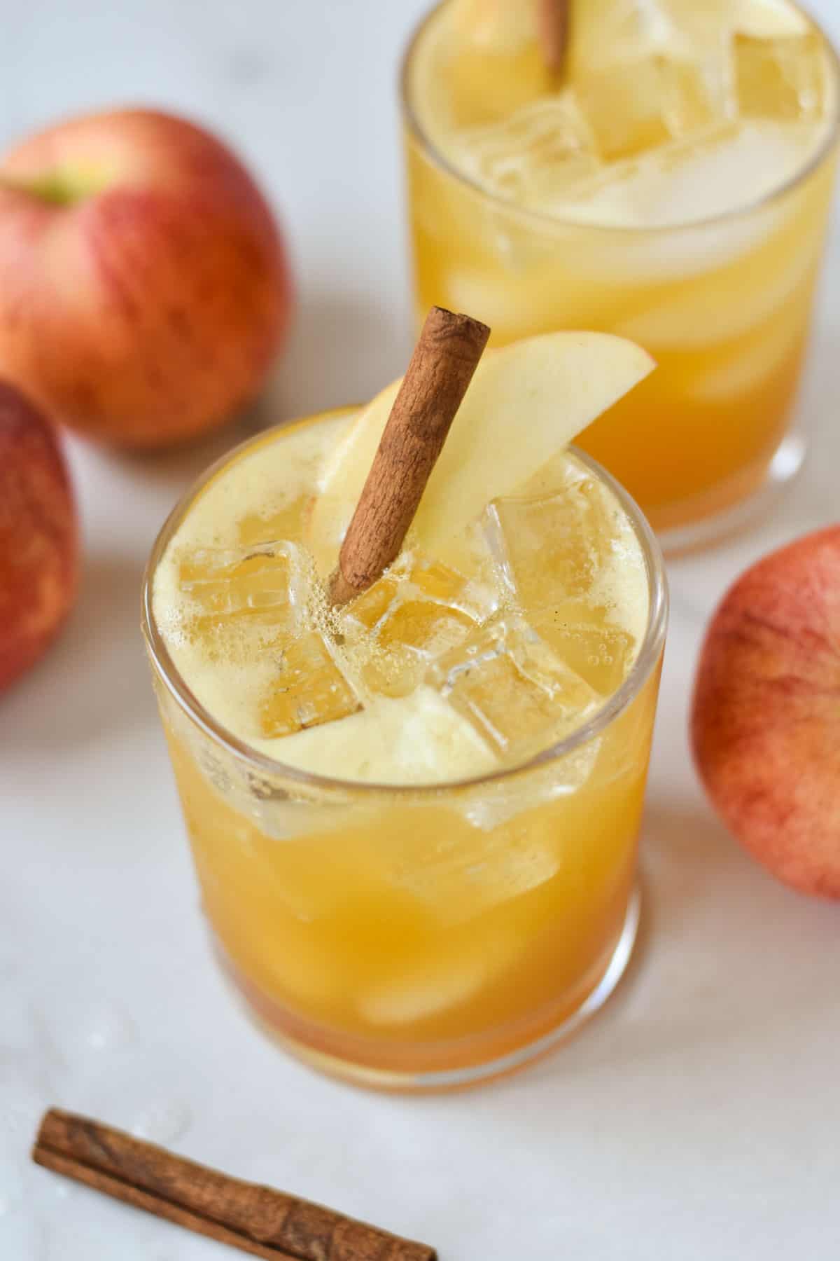 A glass with apple cider and ice, arranged around fresh apples and cinnamon sticks.