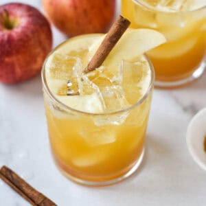 An apple cider mocktail on a white background with cinnamon, apples, and turmeric in a small white bowl.