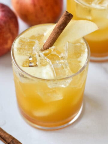 An apple cider mocktail on a white background with cinnamon, apples, and turmeric in a small white bowl.