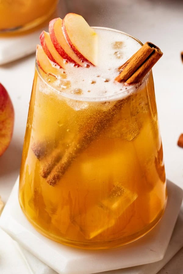 A fall mocktail topped with cinnamon sticks and apples in a clear glass with ice.