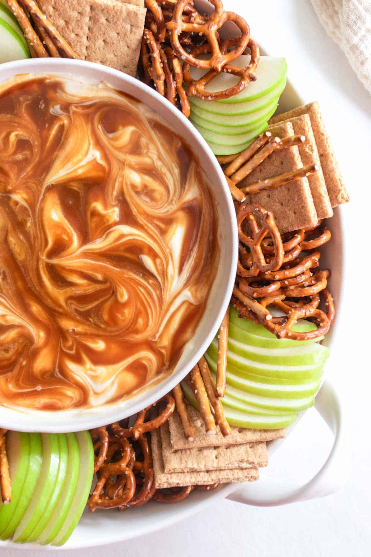 Swirled caramel on cream cheese on a plate with green apples, pretzels, and graham crackers.