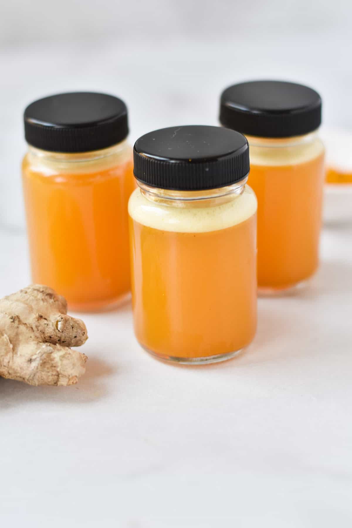 Three turmeric ginger juice bottles next to a knob of fresh ginger.