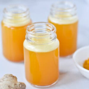 Three ginger turmeric shots in glass bottles next to fresh ginger root and turmeric.