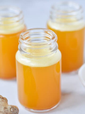 Three ginger turmeric shots in glass bottles next to fresh ginger root and turmeric.