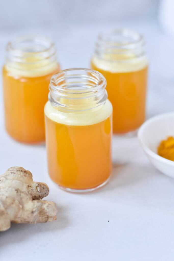 Three glass jars with an orange turmeric juice next to fresh ginger and ground turmeric.