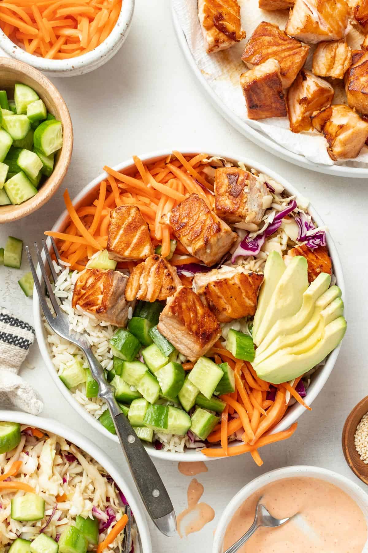 Seared salmon in a bowl with rice, avocado, carrots, slaw, and a small bowl of spicy mayo on the side.