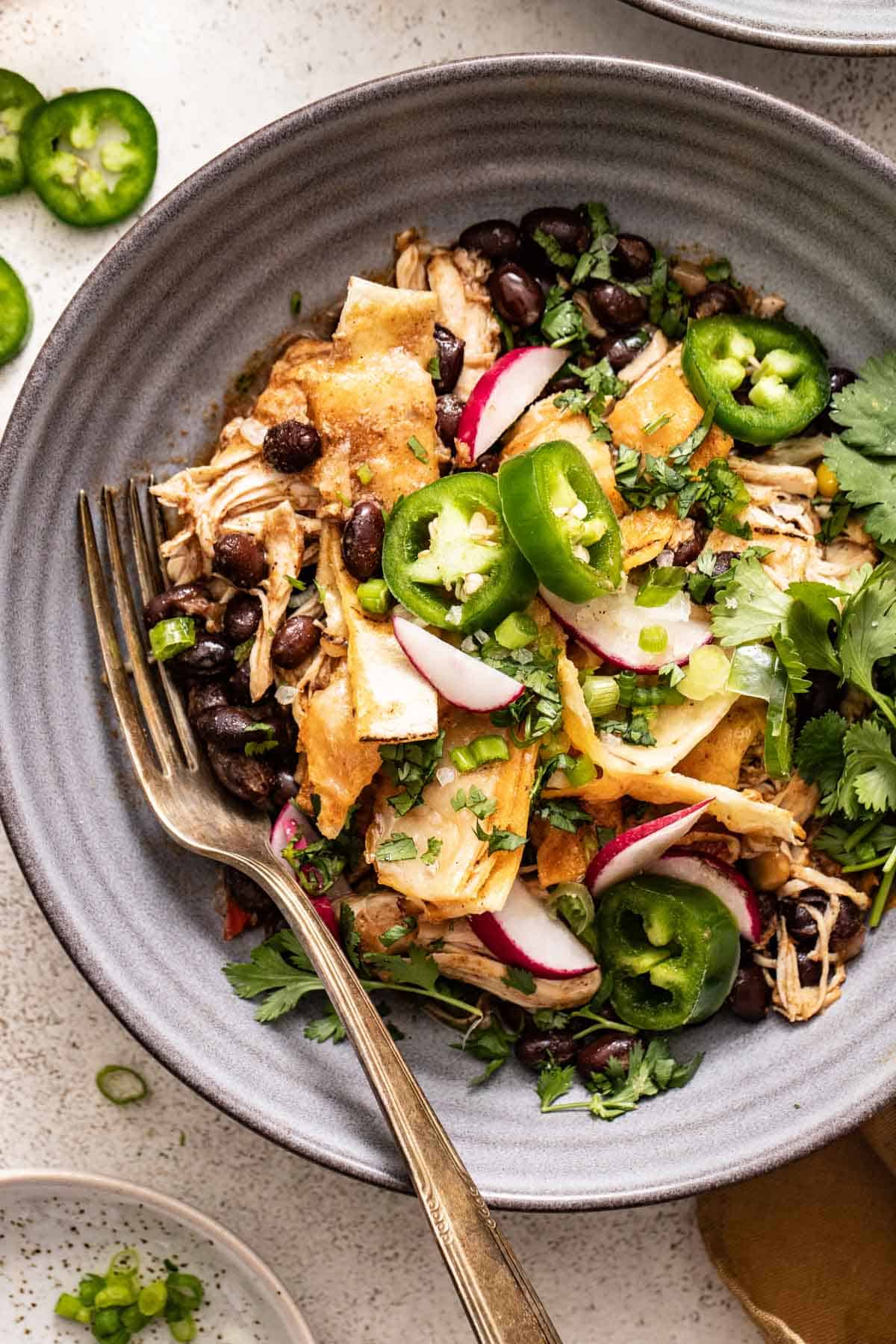 A bowl with chicken enchilada casserole topped with sliced jalapenos, next to a fork.