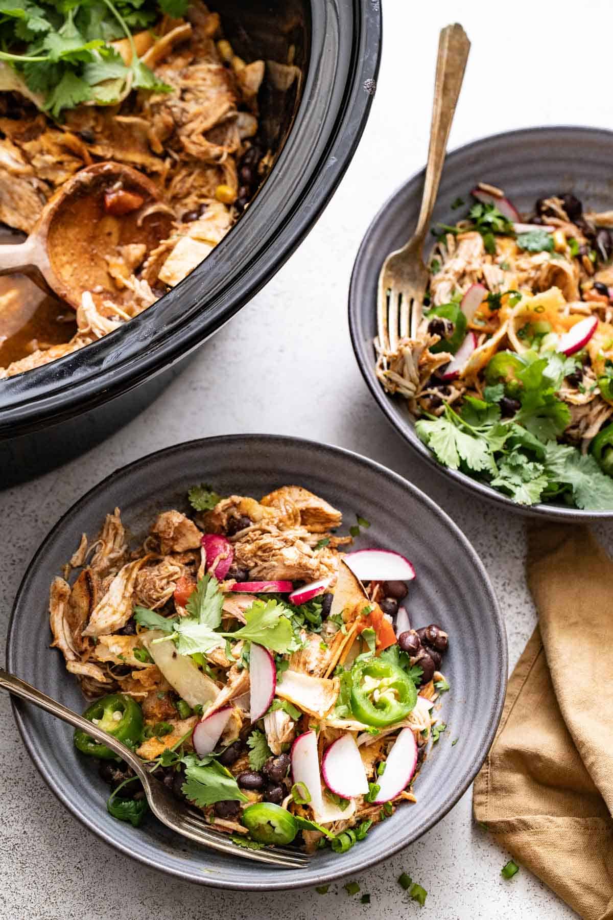 Two bowl of chicken enchilada filling next to a slow cooker.