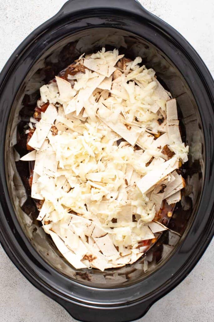 Chicken enchiladas in a slow cooker being topped with shredded cheese.