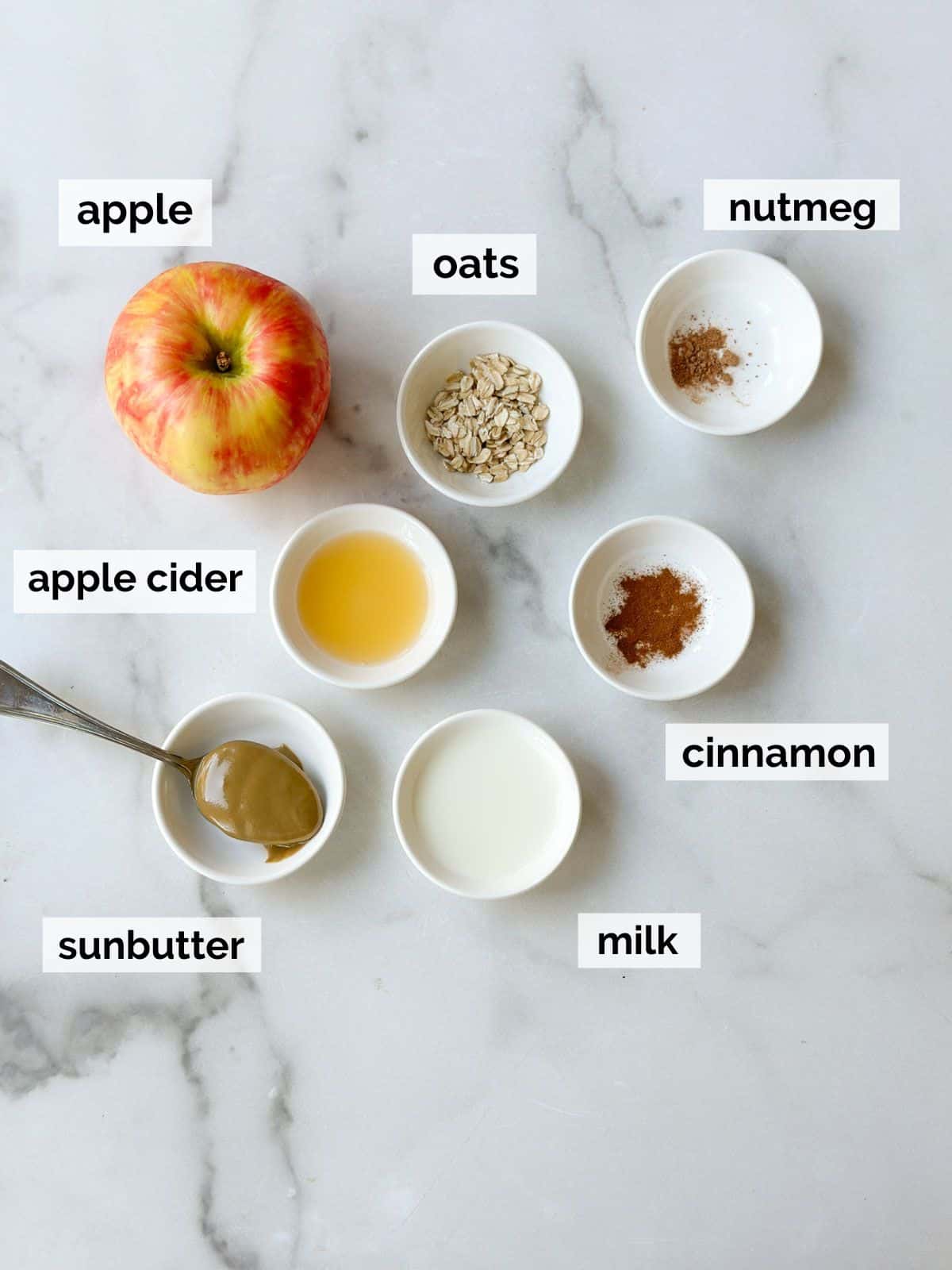 Ingredients containing apples, oats, and apple cider on a marble background.