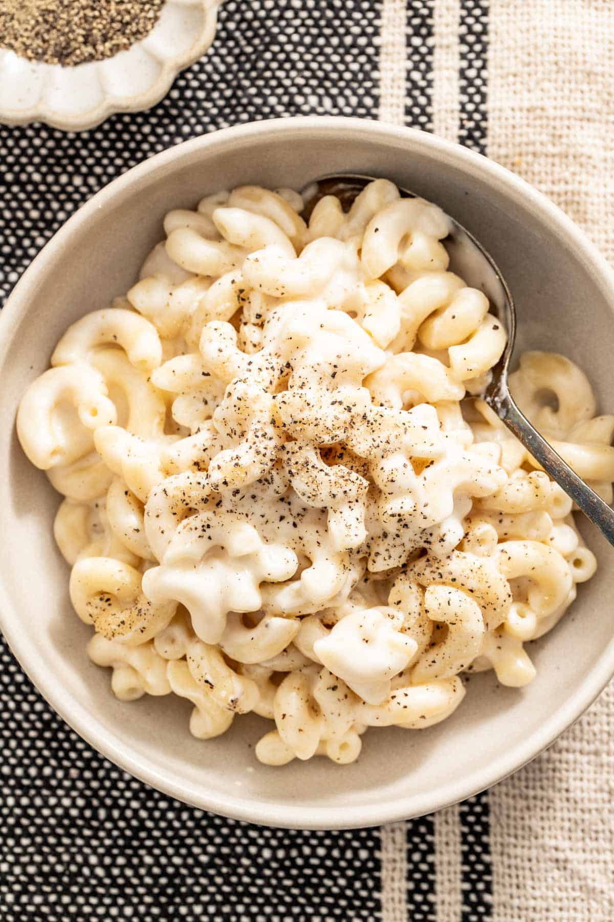 Mac and cheese in a creamy sauce in a white bowl with a spoon and black pepper on top.