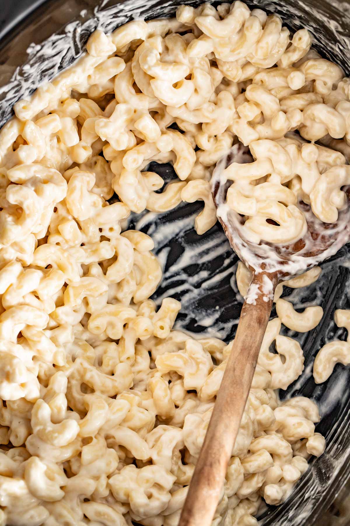 A wood spoon in a slow cooker with mac and cheese and cream cheese on top.