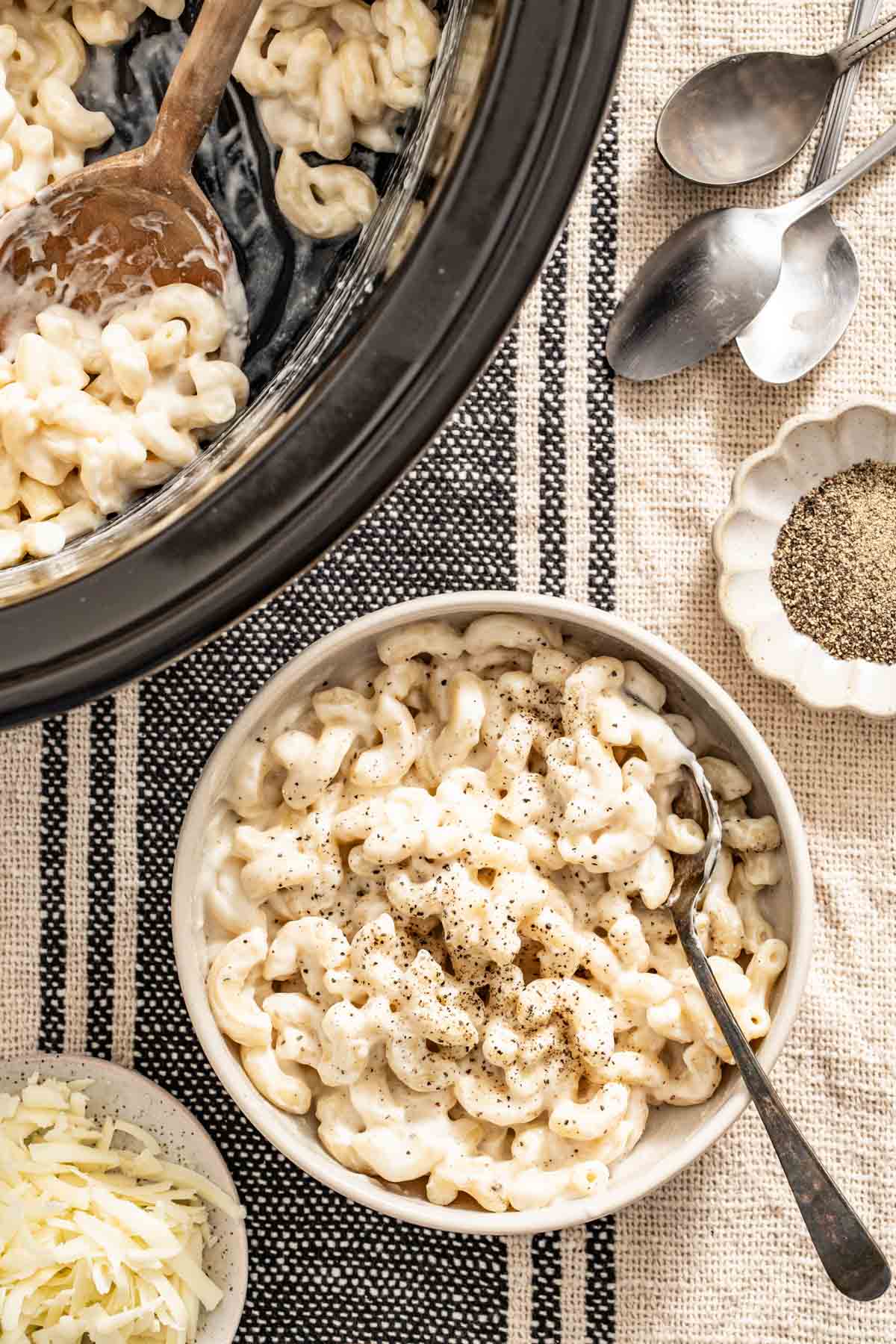 A bowl of white mac and cheese next to shredded cheese and a slow cooker with extra macaroni.