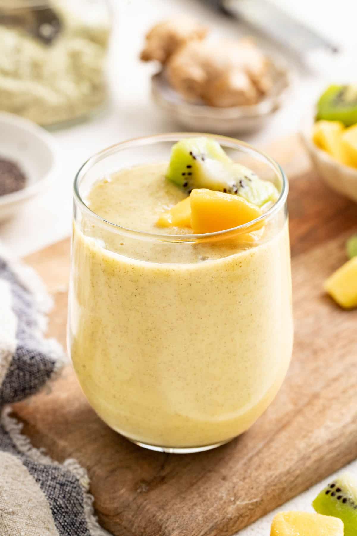 A yellow ginger smoothie topped with mango and kiwi on a wood board.