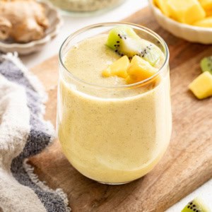 A yellow ginger smoothie topped with mango and kiwi on a wood board.