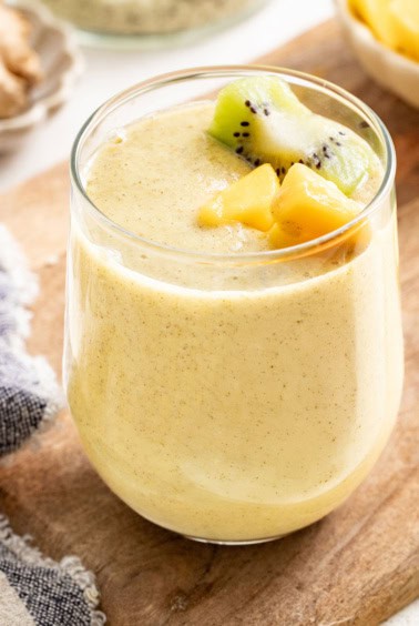 A yellow ginger smoothie topped with mango and kiwi on a wood board.