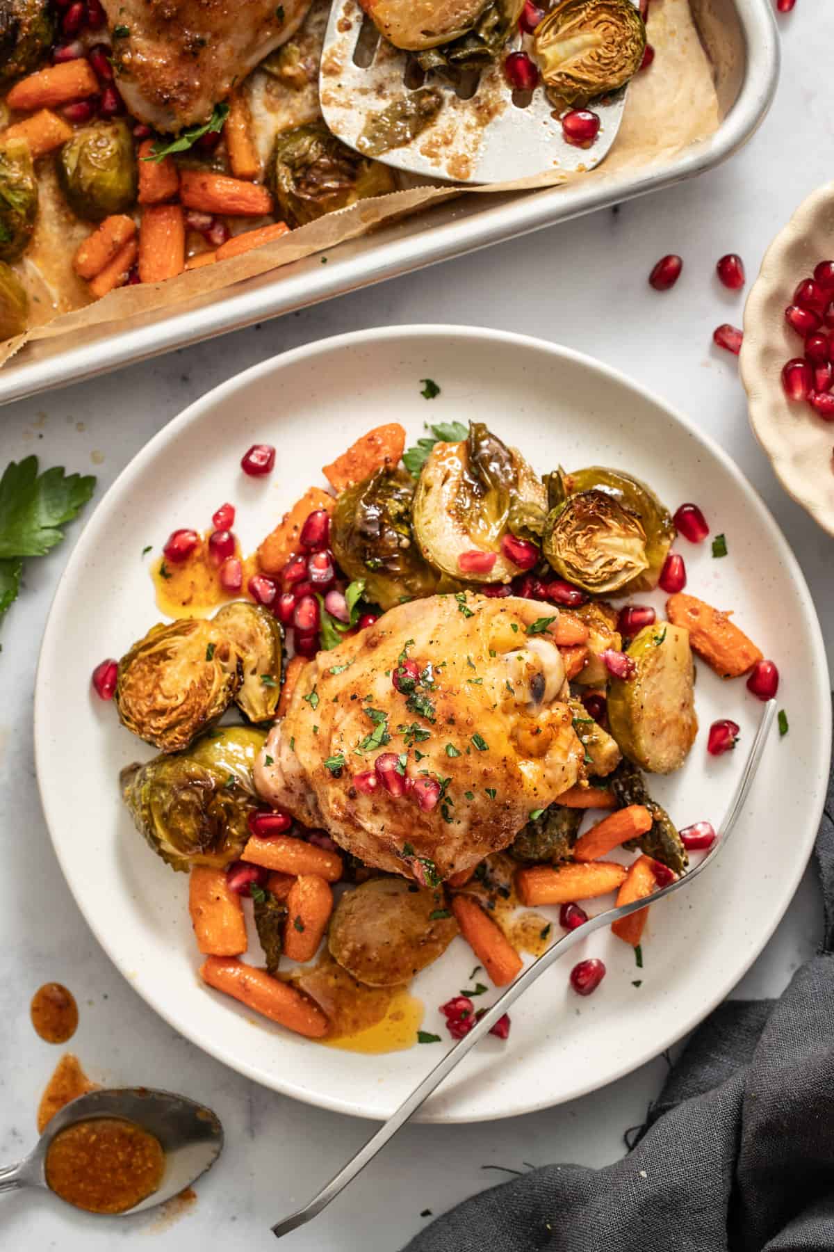 A plate with roasted chicken thighs topped with pomegranate seeds, next to brussels sprouts and carrots.