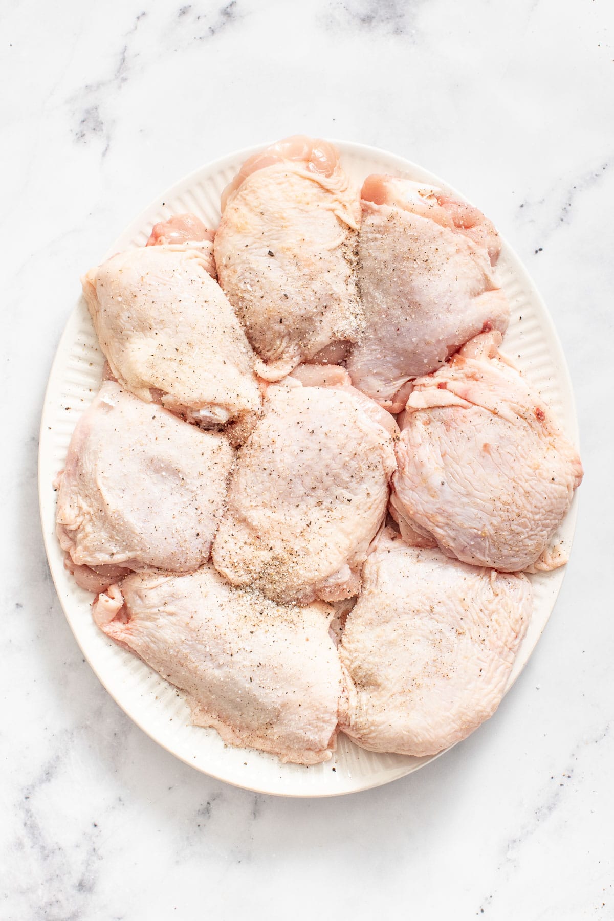 Bone in skin on chicken thighs seasoned with salt and pepper on a white plate. 