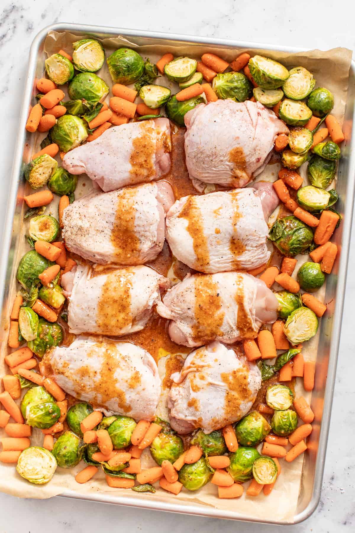 Maple dijon sauce poured on top of unbaked chicken thighs with brussels and carrots on a sheet pan.