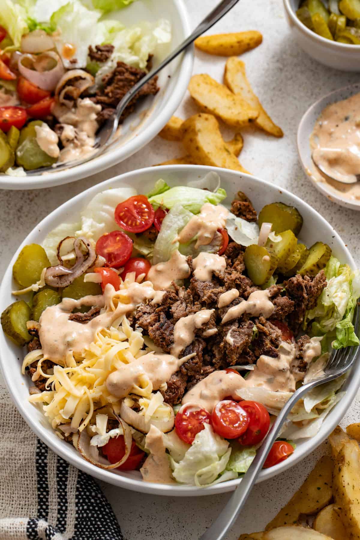 Burger bowls topped with a homemade big mac sauce next to a bowl of sauce and roasted potato wedges.
