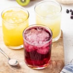 3 flavors of homemade gatorade in glasses on a wood table next to a spoon with salt.