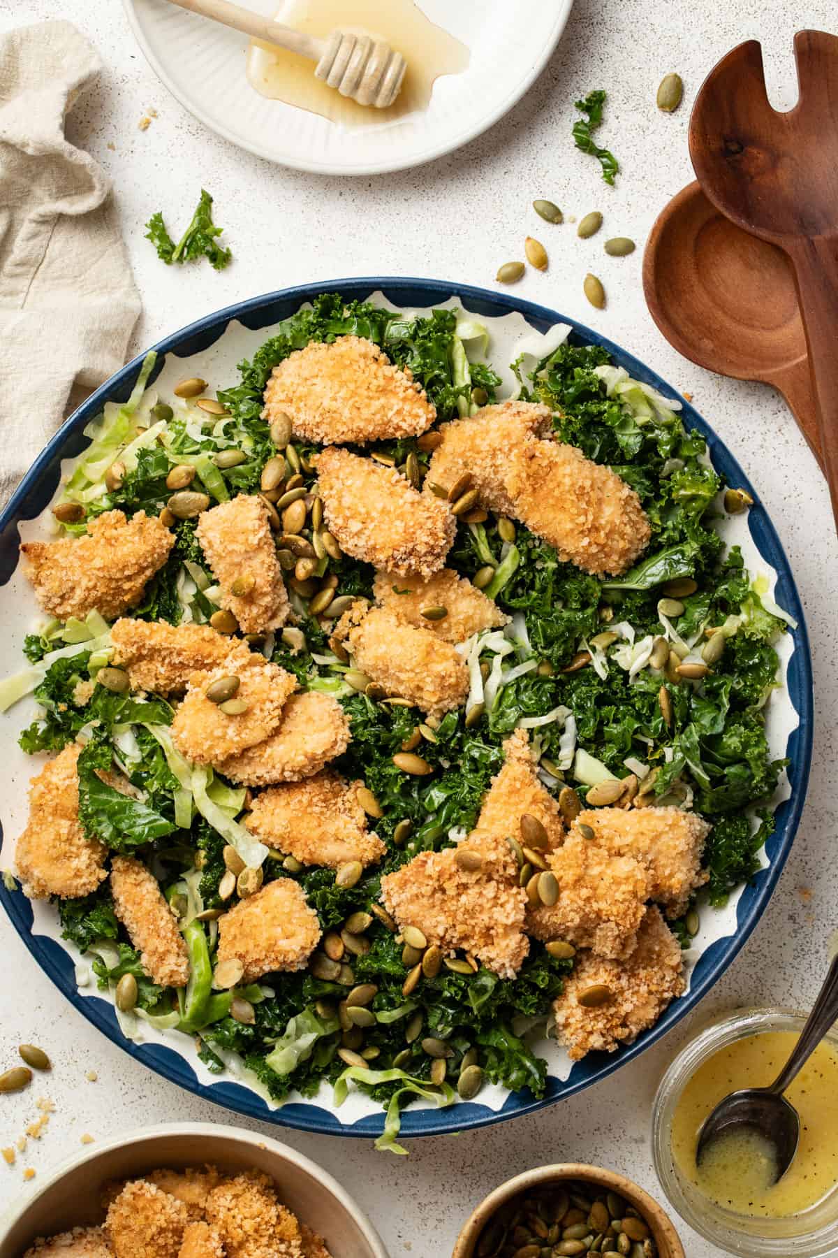 A kale salad in a large bowl next to dressing, chicken nuggets, and seeds. 