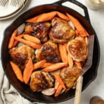 Crispy bone in chicken thighs and carrots in a cast iron pan with a spatula after being seared.