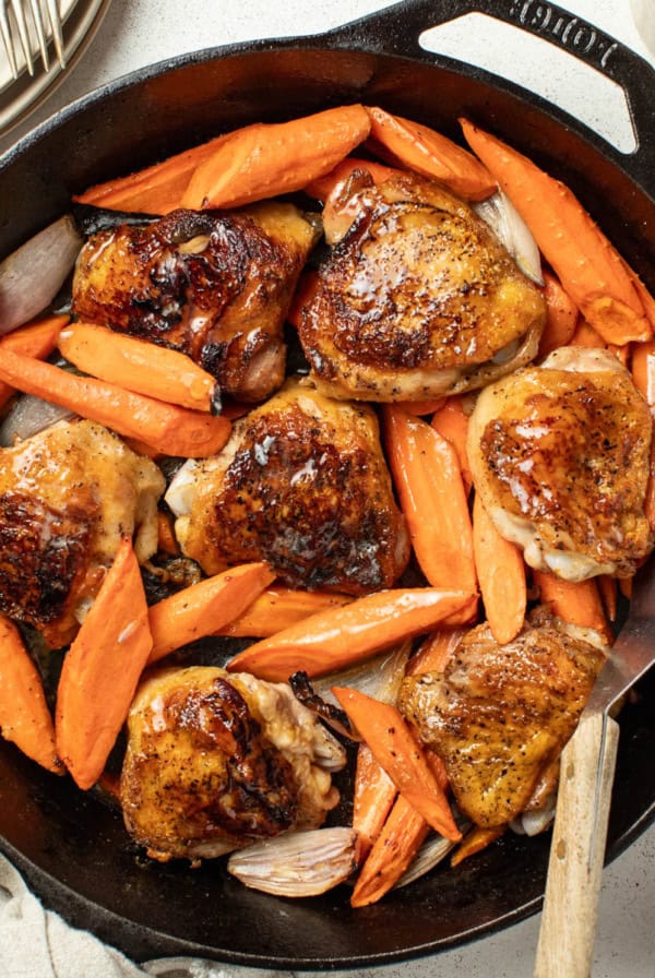 Crispy bone in chicken thighs and carrots in a cast iron pan with a spatula after being seared.