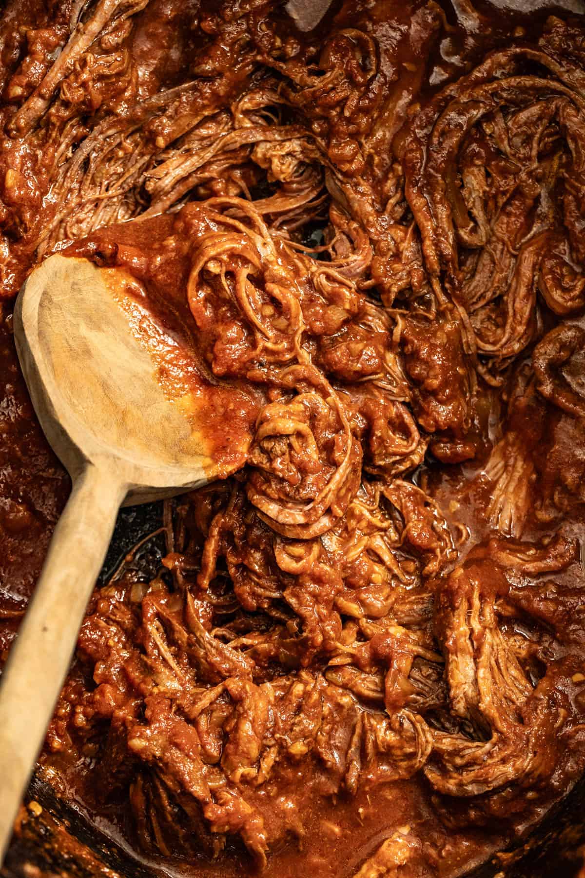 A wood spoon scooping shredded flank steak in a slow cooker.
