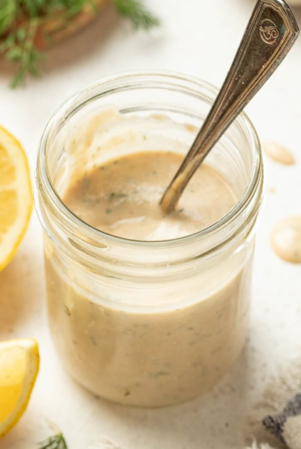 Tahini sauce in a mason jar with a spoon next to lemons and dill.