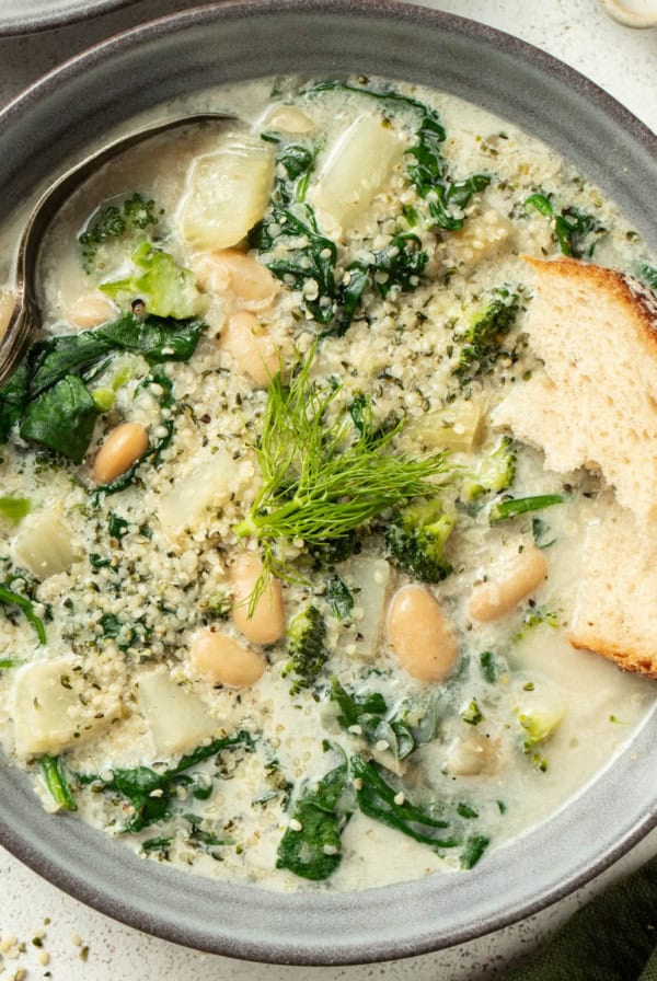 A creamy white bean soup with veggies and crusty bread dipping into it.