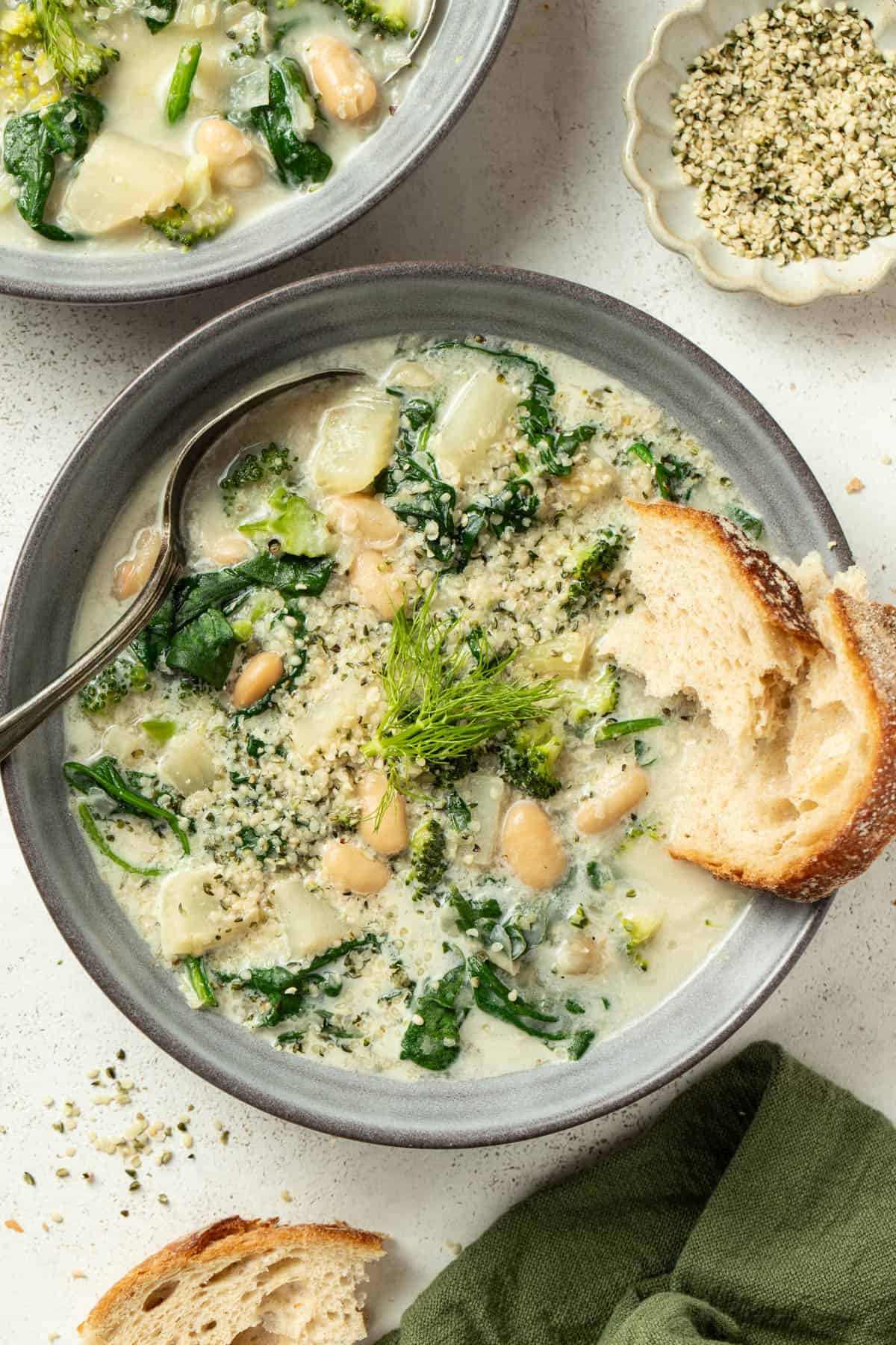 A creamy white bean soup with veggies and crusty bread dipping into it next to a spoon.