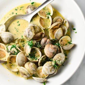 Steamed clams in a creamy broth, topped with parsley in a giant white bowl with a stainless steel spoon dipping in.