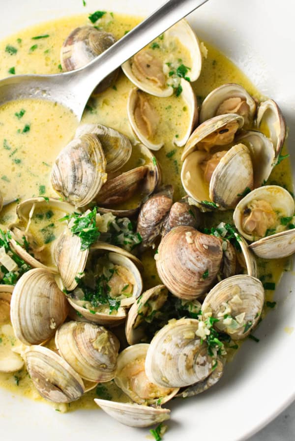 Steamed clams in a creamy broth, topped with parsley in a giant white bowl with a stainless steel spoon dipping in.