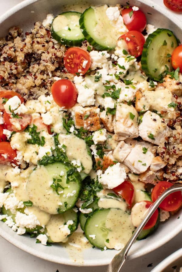 A chicken quinoa bowl with tomatoes, cucumber, and feta and a Mediterranean dressing drizzled on top.