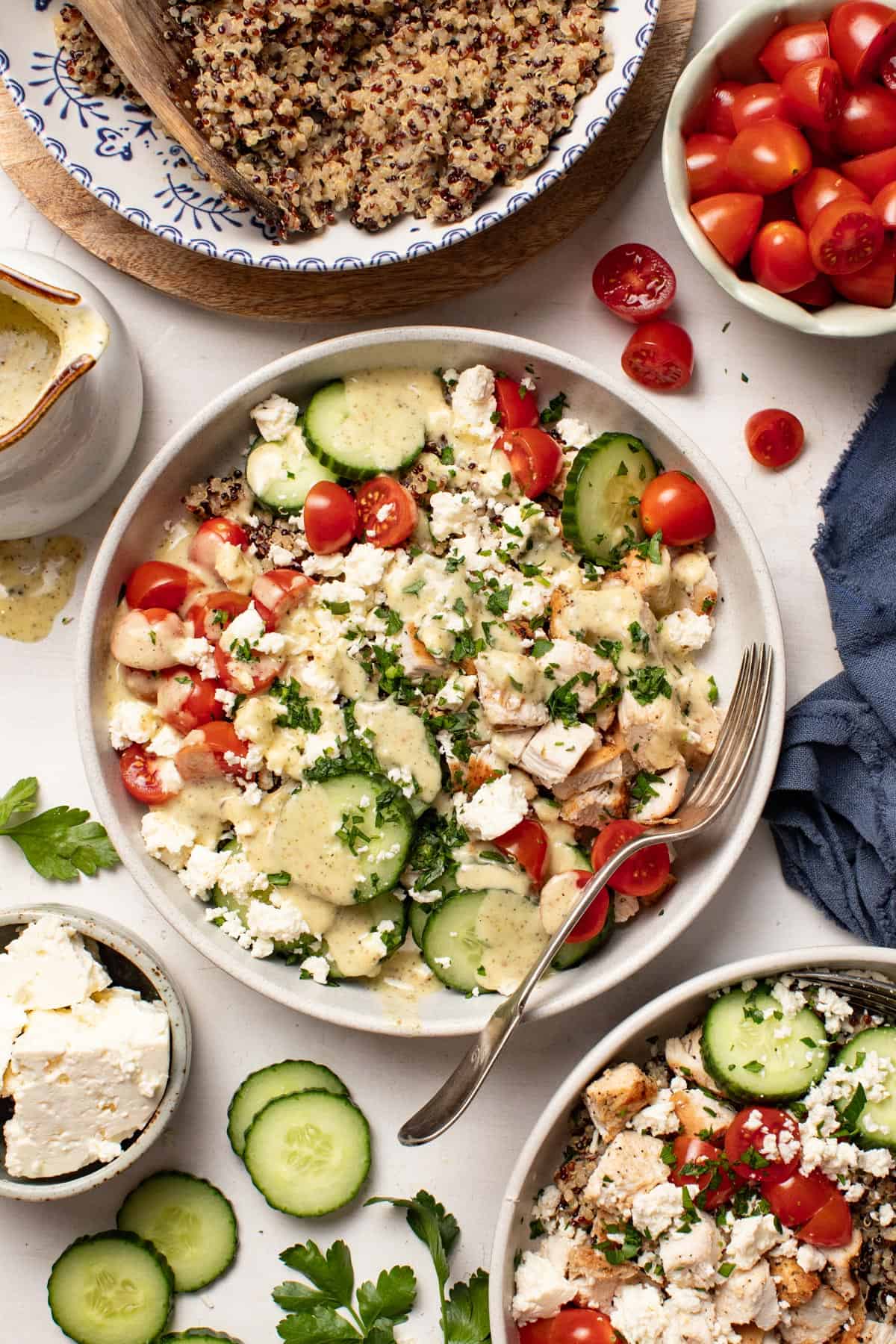 A Mediterranean chicken quinoa bowl next to a bowl with tomatoes and cucumbers, a bowl with feta, and a big bowl of cooked quinoa to the side.