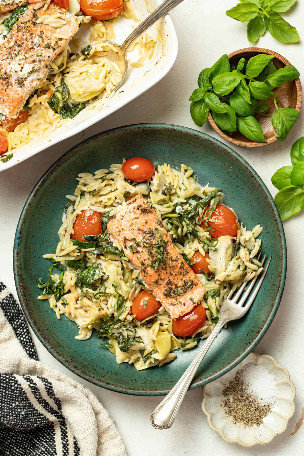 Salmon and orzo in a bowl next to a baking dish with more baked salmon and basil.