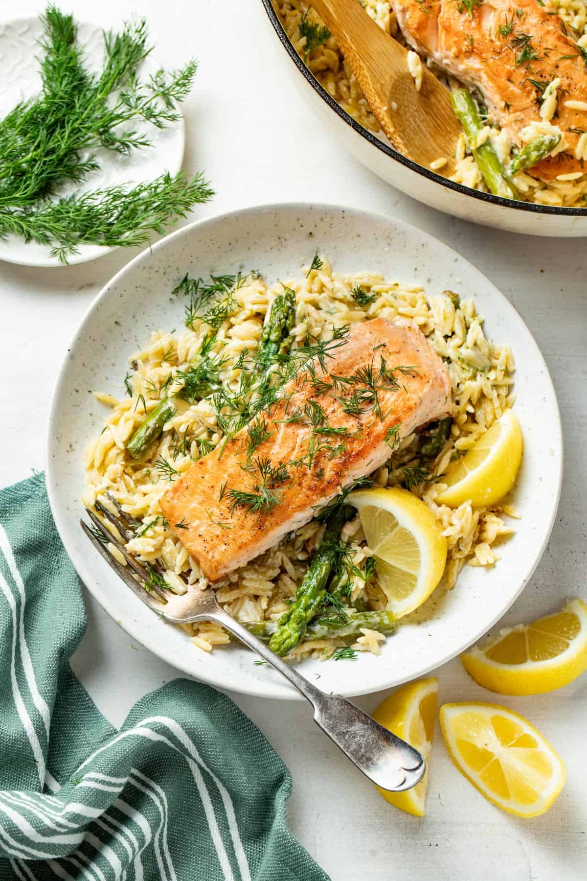 A salmon fillet on top of creamy lemon orzo on a plate with a fork and lemon slices next to it.