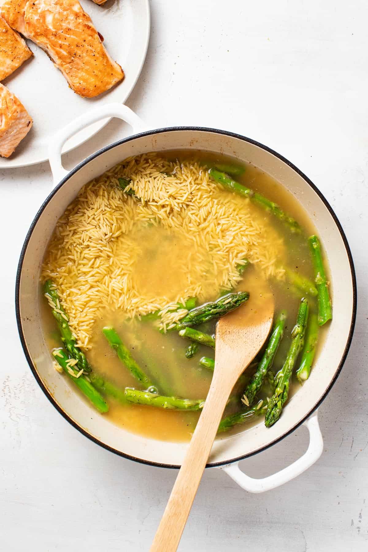 Adding orzo and broth to a pan with fresh asparagus.