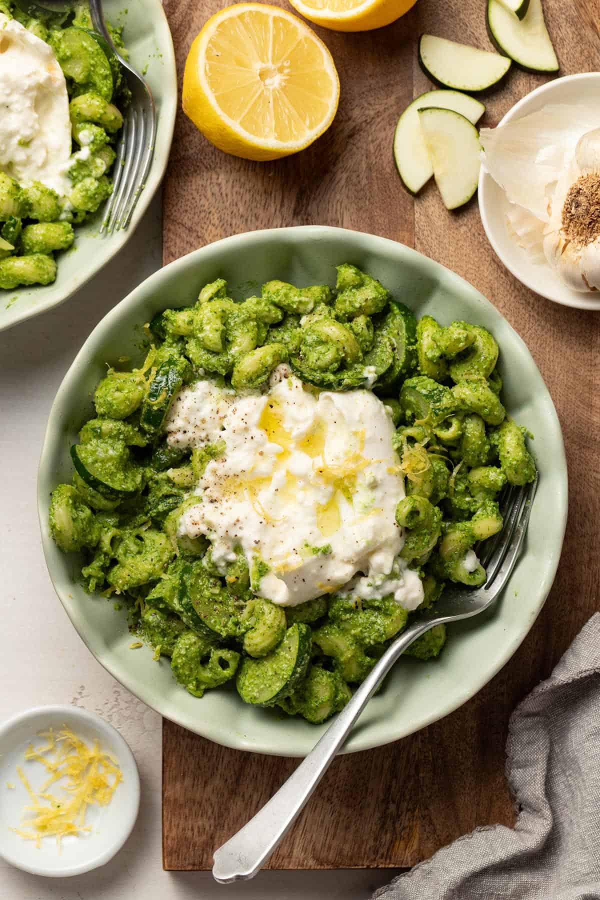 A bowl of burrata pasta topped with zucchini, olive oil, and lemon zest next to a bowl with zucchini wedges.